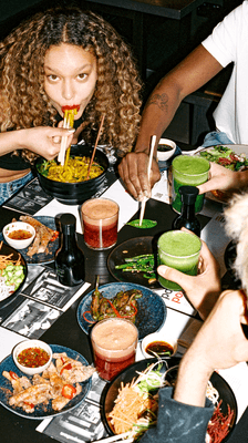 a woman eating noodles with chopsticks sat at a table of food and drink