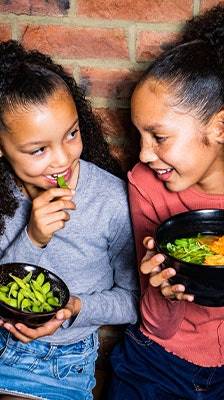 2 girls hold black bowls with edamame and ramen