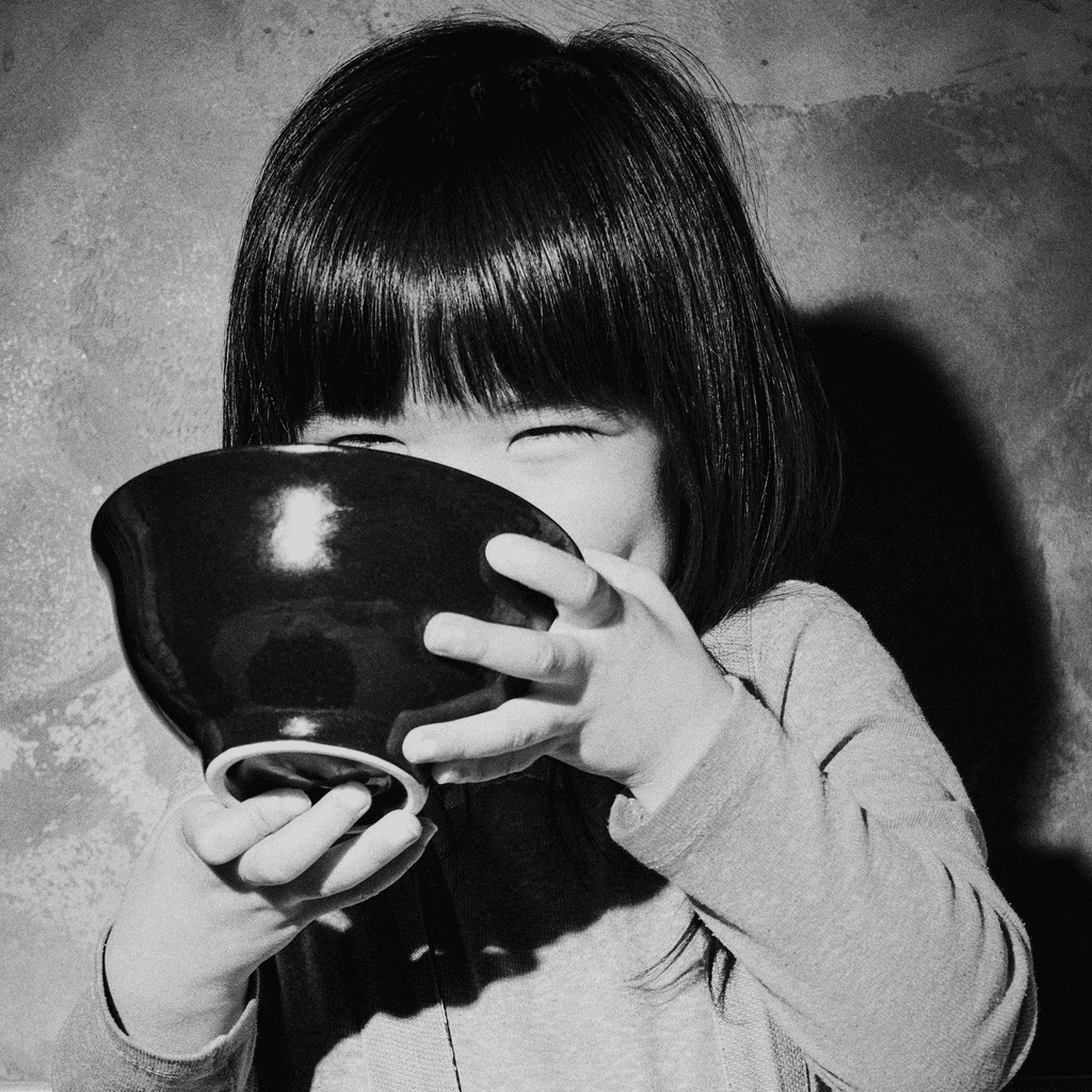 a little girl drinks ramen from a black bowl