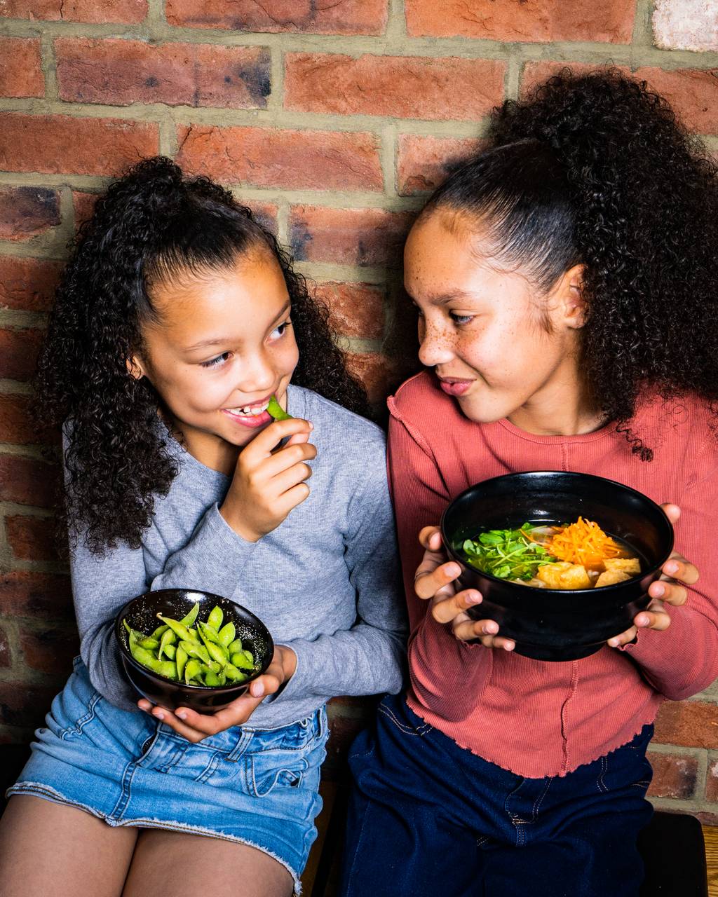 2 girls hold black bowls with edamame and ramen